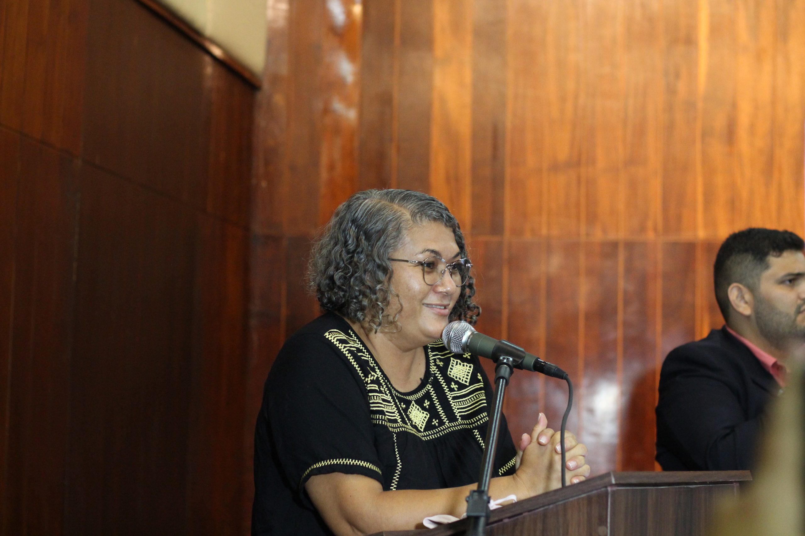 Marinalva Santana representante do grupo Matizes discursa em coloca as duas mãos em cima do púlpito. A mulher é negra, tem cabelos cacheados, usa óculos e blusa preta
