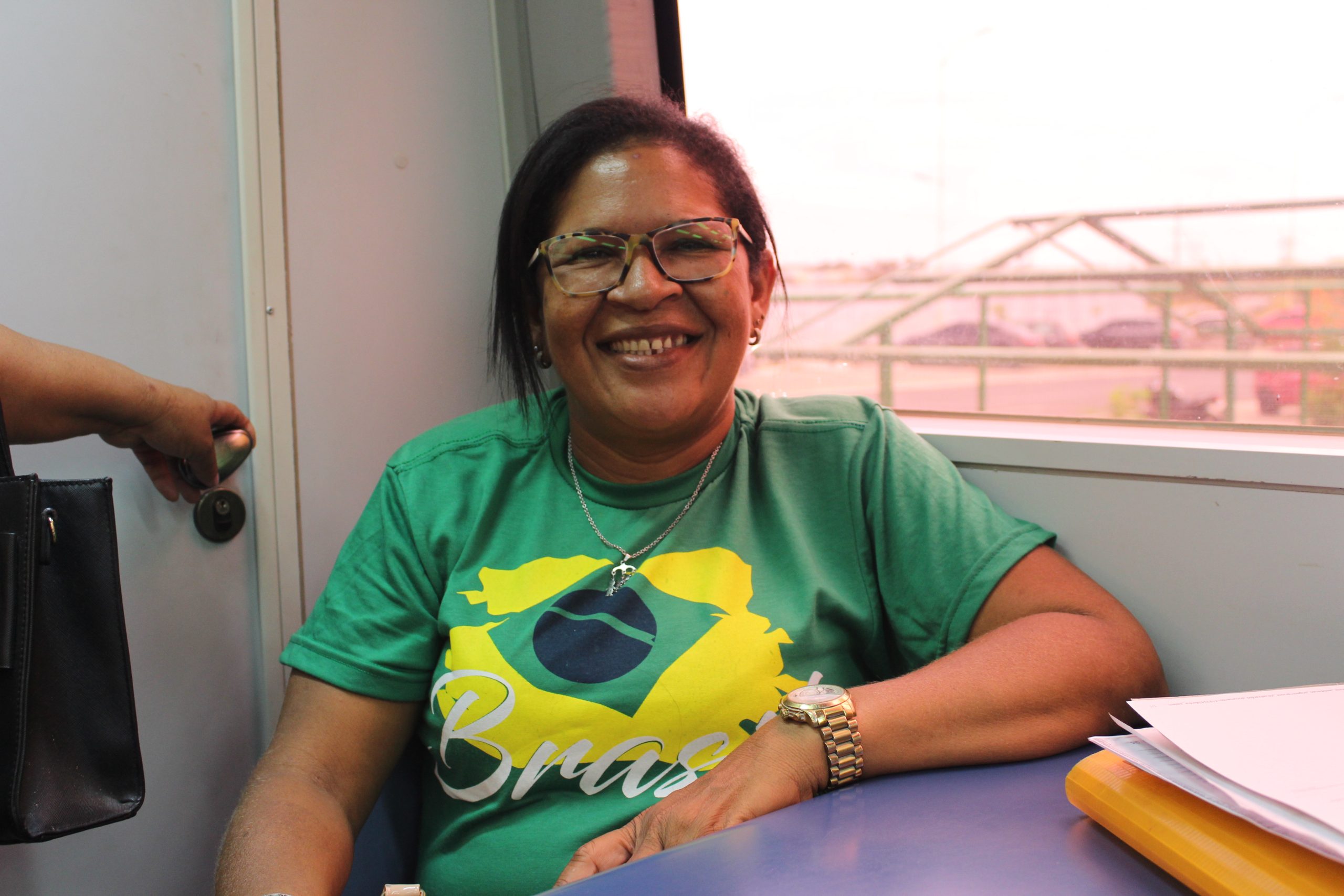 Raquel Batista esta sentada sorrindo para foto. Mulher de pele negra, usa óculos, cabelo liso, vestido em uma blusa com estada a bandeira do Brasil, nas cores verde, amarela, azul e branco. 
