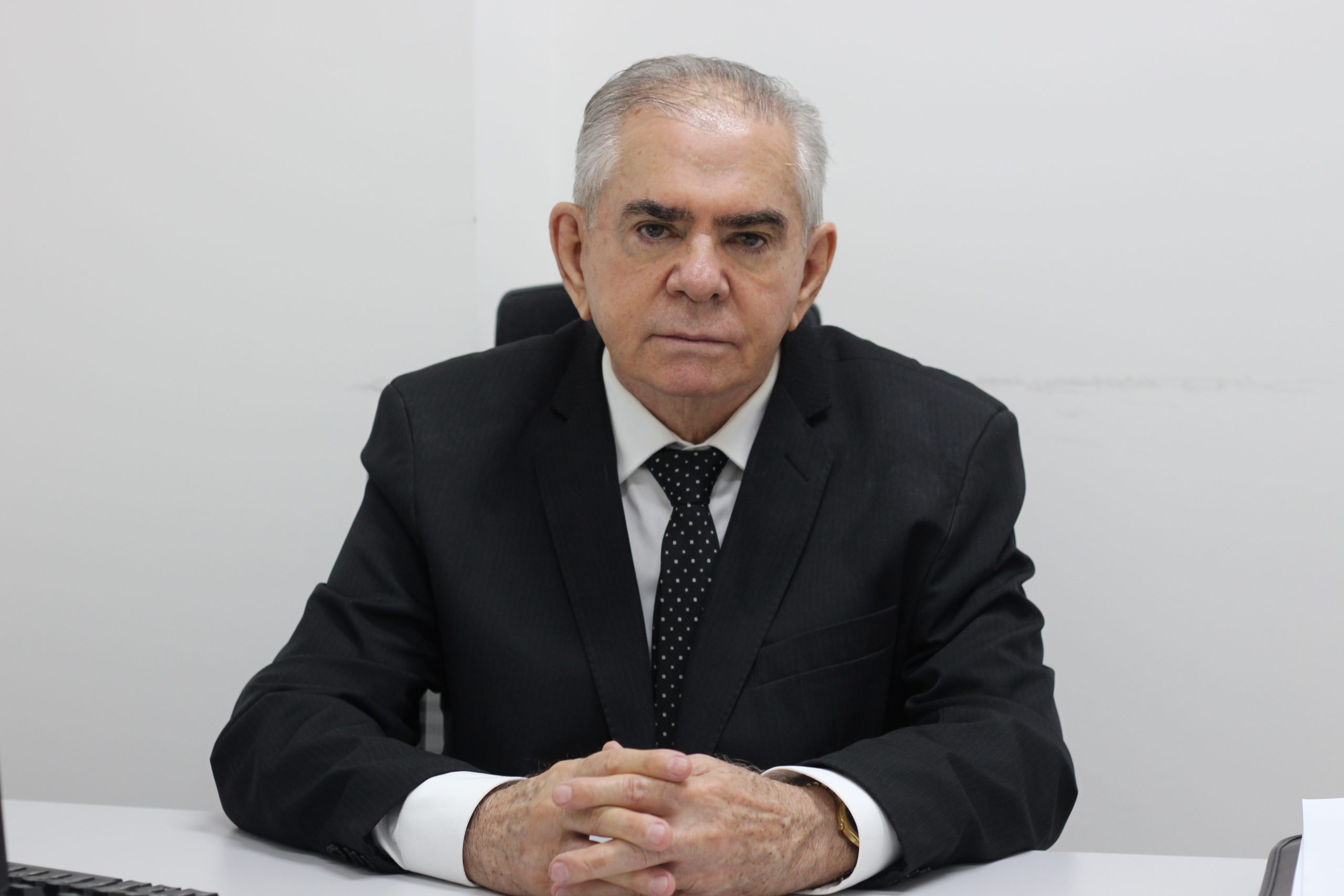 Médico Leopoldino Ferreira Filho esta sentado em uma cadeira com as mãos em cima da mesa. Usa gravata com bolinhas branca, tem cabelos grisalhos. 