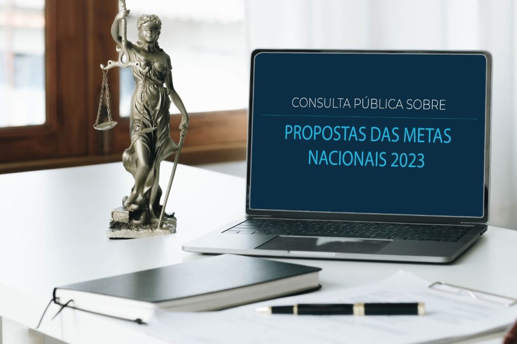 Na imagem: uma mesa branca com uma caderneta, uma pequena escultura da deusa Artêmis representeando a justiça e um notebook aberto, inscrito na tela "Consulta pública sobre propostas das metas nacionais 2023"