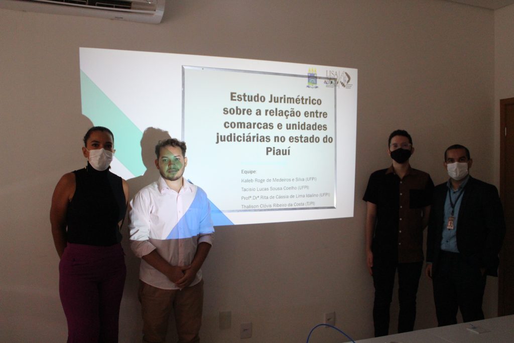 Na imagem: a professora, o representante do tribunal e os dois alunos estão em frente a uma apresentação de slide, com o tiútulo "Estudo jurimétrico sobre a relação entre comarcas e unidades judiciárias no estado do Piauí"