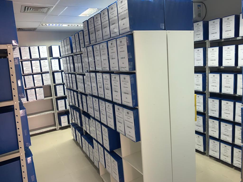 Na imagem: caixas de arquivos azuis, em estantes brancas, em uma sala branca.