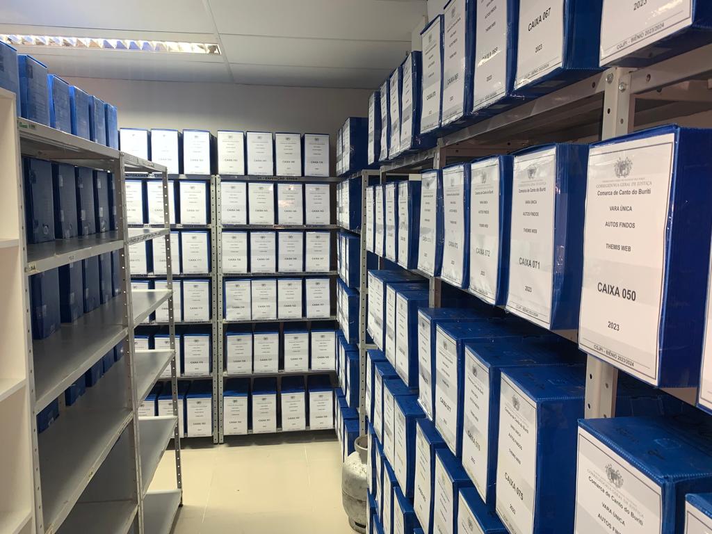 Na imagem: caixas de arquivos azuis, em estantes brancas, em uma sala branca.
